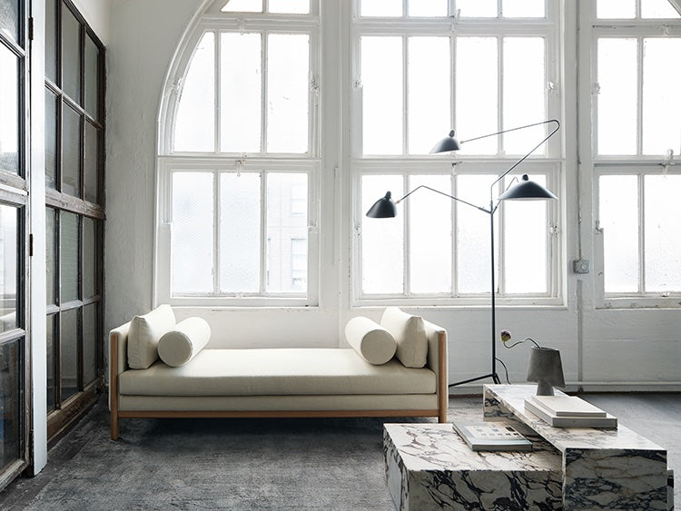 A minimalist living space features a modern white sofa with cylindrical cushions, a marble coffee table with books on top, and a contemporary floor lamp. Large arched windows allow abundant natural light, highlighting the room's bright and airy atmosphere.