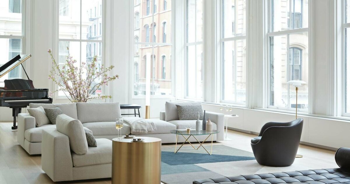 A modern living room with large windows, light-colored sofas, a black armchair, a glass coffee table, and a grand piano. The room is decorated with a minimalistic style, featuring a vase of flowers and a golden side table. Outside, tall buildings are visible.