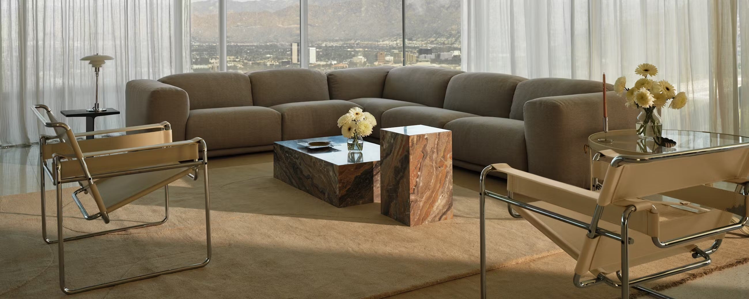 A modern living room featuring a large beige sectional sofa, two beige and chrome lounge chairs, a stone coffee table, and a matching side table. The space is decorated with vases of white flowers and has floor-to-ceiling windows offering a scenic mountain view.