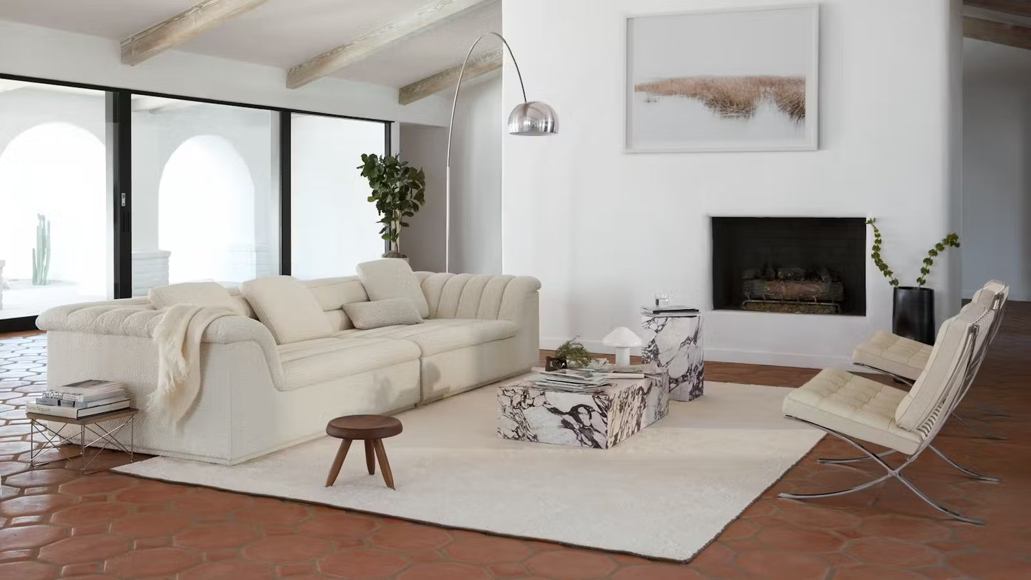 A stylish living room features a white plush sofa, a marble-patterned coffee table, a modern floor lamp, a cozy fireplace, and a contemporary chair on a light rug. Large glass doors open to a bright outdoor space, and a potted plant adds a touch of greenery.