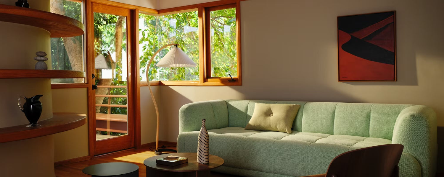 A cozy living room featuring a green sofa with a beige pillow, a round wooden coffee table with a striped vase, a tall floor lamp, wooden shelves with decorative items, and a large window allowing natural light from a leafy outdoor scene. A modern painting adorns the wall.