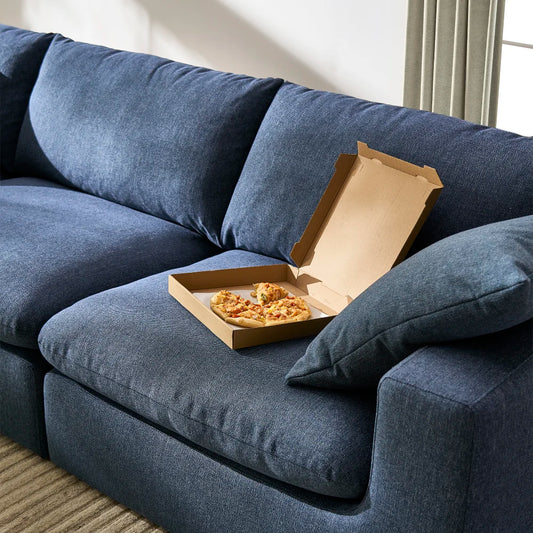 A dark blue sofa with a cardboard pizza box containing a few slices of pizza placed on the seat cushion. Sunlight filters through nearby curtains, creating a warm atmosphere in the room. A beige rug partially visible on the floor.