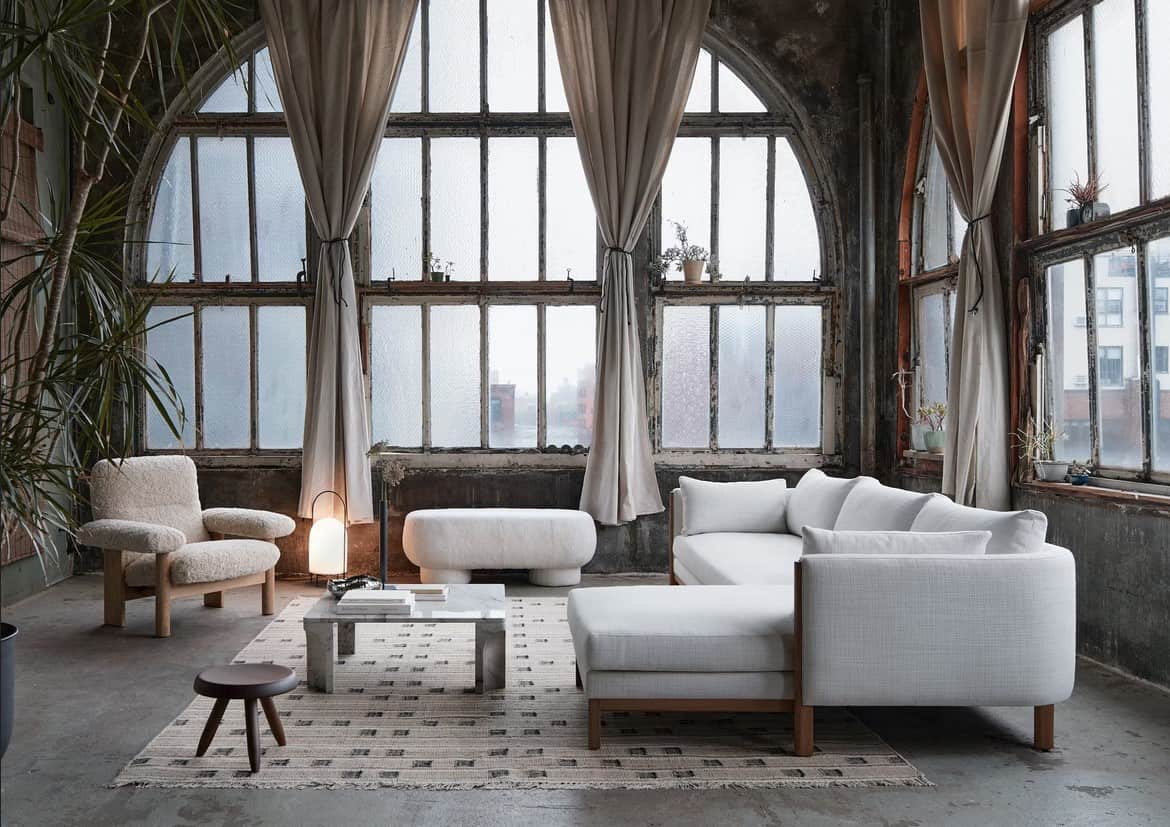 A spacious room with large arched windows covered with sheer white curtains. The room features a white sectional sofa, a matching ottoman, a textured armchair, a low coffee table, a patterned rug, and a small wooden stool. Plants and a floor lamp add warmth.