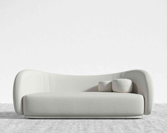 The Diane Sofa is a modern, minimalist piece with smooth, curved edges and light gray upholstery. It includes two square cushions and sits on a light gray carpet against a plain white wall.