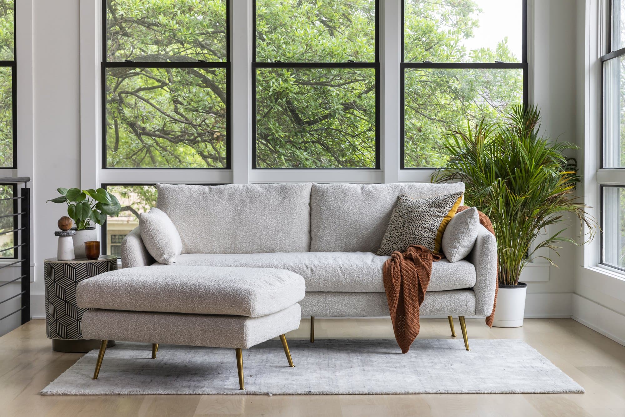 A modern living room features a light-gray sofa with chaise lounge, adorned with a rust-colored throw blanket and a mix of patterned cushions. The sofa sits on a gray rug beside tall windows with a view of lush greenery. Nearby are a plant and a small side table.