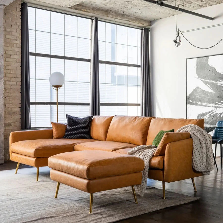 A modern living room featuring a brown leather sectional sofa with a matching ottoman. The sofa is adorned with a gray throw blanket and green pillow. Large windows, gray curtains, and a floor lamp with a round white shade complement the space. A monochrome abstract painting hangs on the wall.