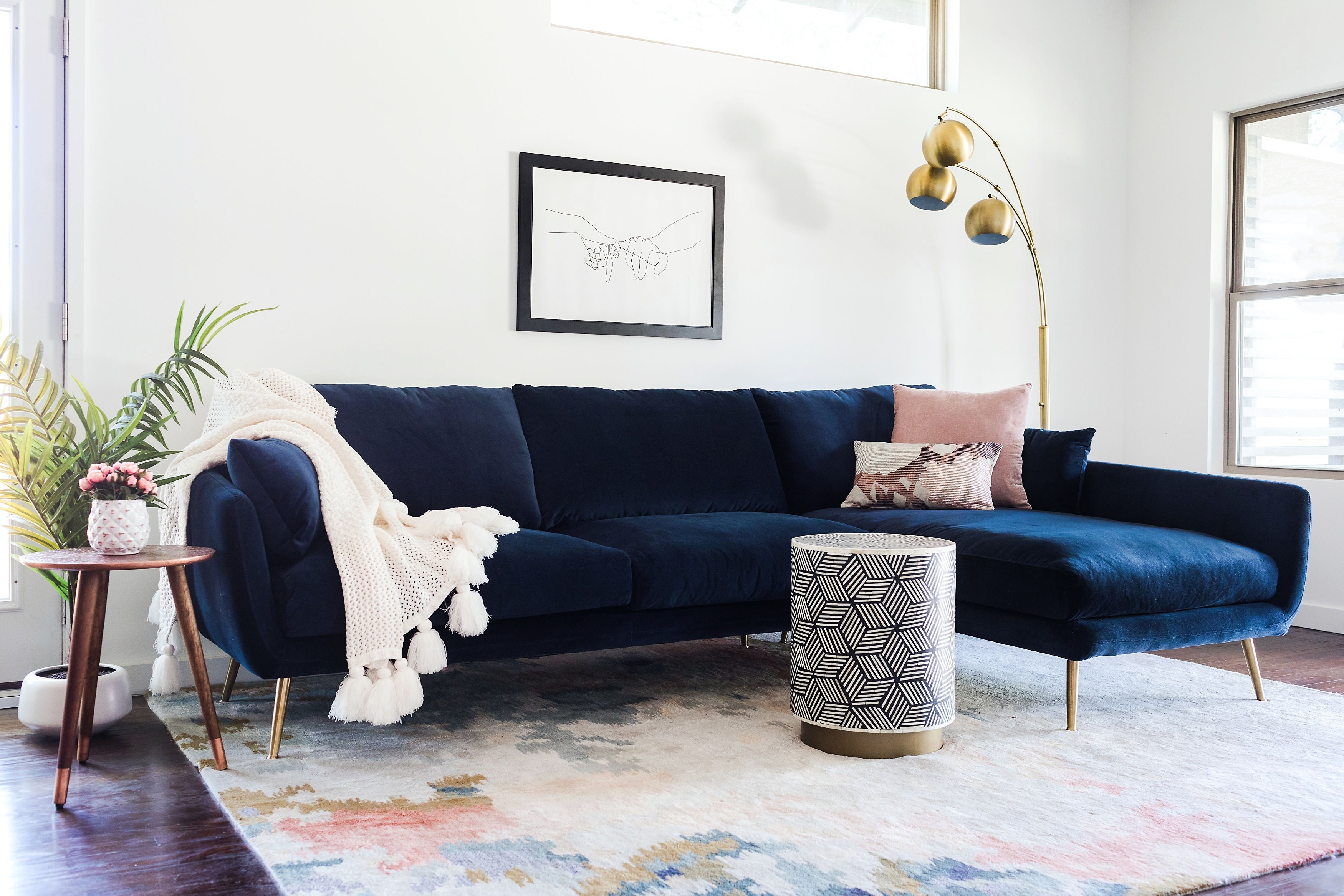 A modern living room features a navy blue sectional sofa with a white fringed throw and pink cushions. A geometric side table sits in front, while a gold floor lamp stands beside. There's a framed line drawing on the wall, and plants add greenery against white walls.