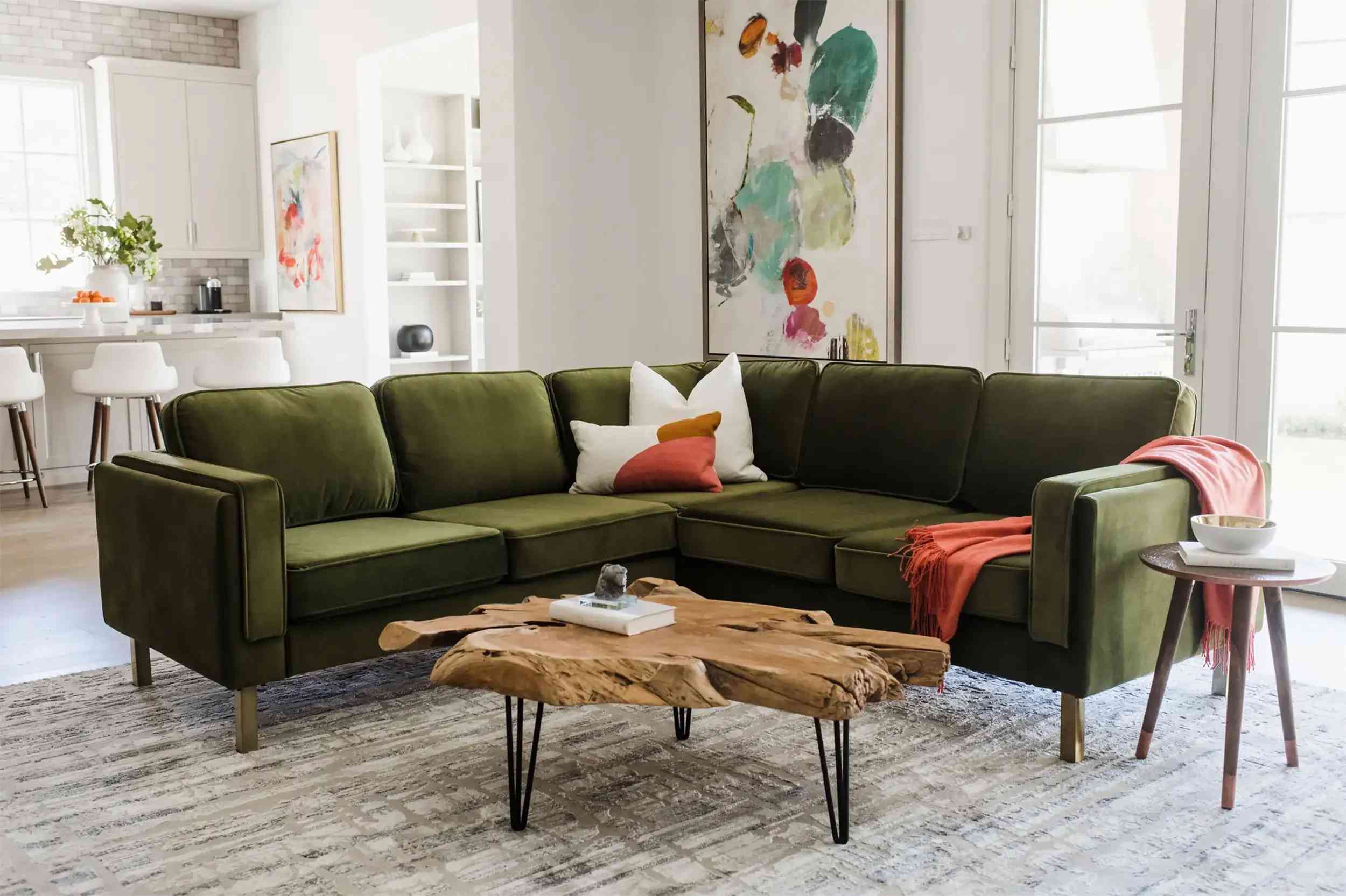 A stylish living room features a green velvet sectional sofa with a red throw and a decorative pillow. In front of the sofa is a unique, wooden coffee table with hairpin legs. A modern abstract painting adorns the wall behind the sofa, and a bright, open kitchen is visible in the background.