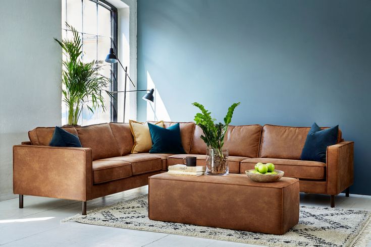 A modern living room with a brown leather sectional sofa adorned with blue and yellow throw pillows. A large ottoman serves as a coffee table, holding a glass vase with green plants and a bowl of green apples. The room has blue walls, large windows, and is accented by a leafy plant in the corner.