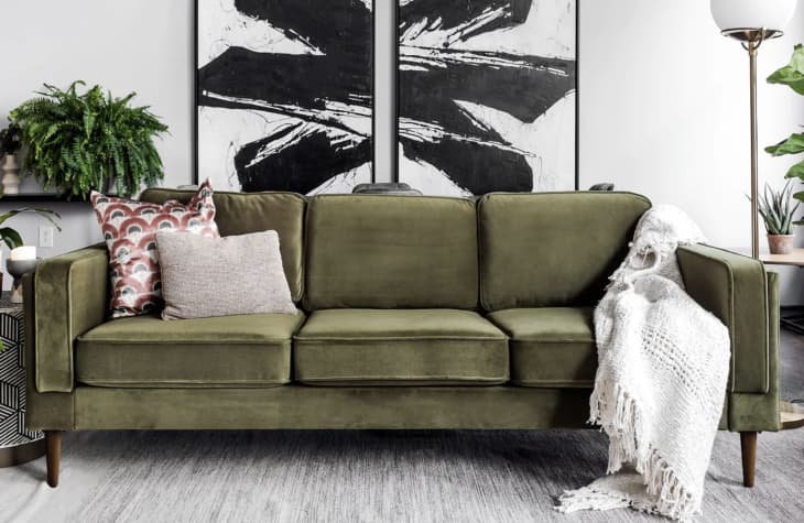 A stylish living room features a green velvet couch adorned with patterned and plain pillows. A white knitted blanket is draped over the armrest. Behind the couch are two black and white abstract paintings. There's a hanging fern and a modern floor lamp in the background.