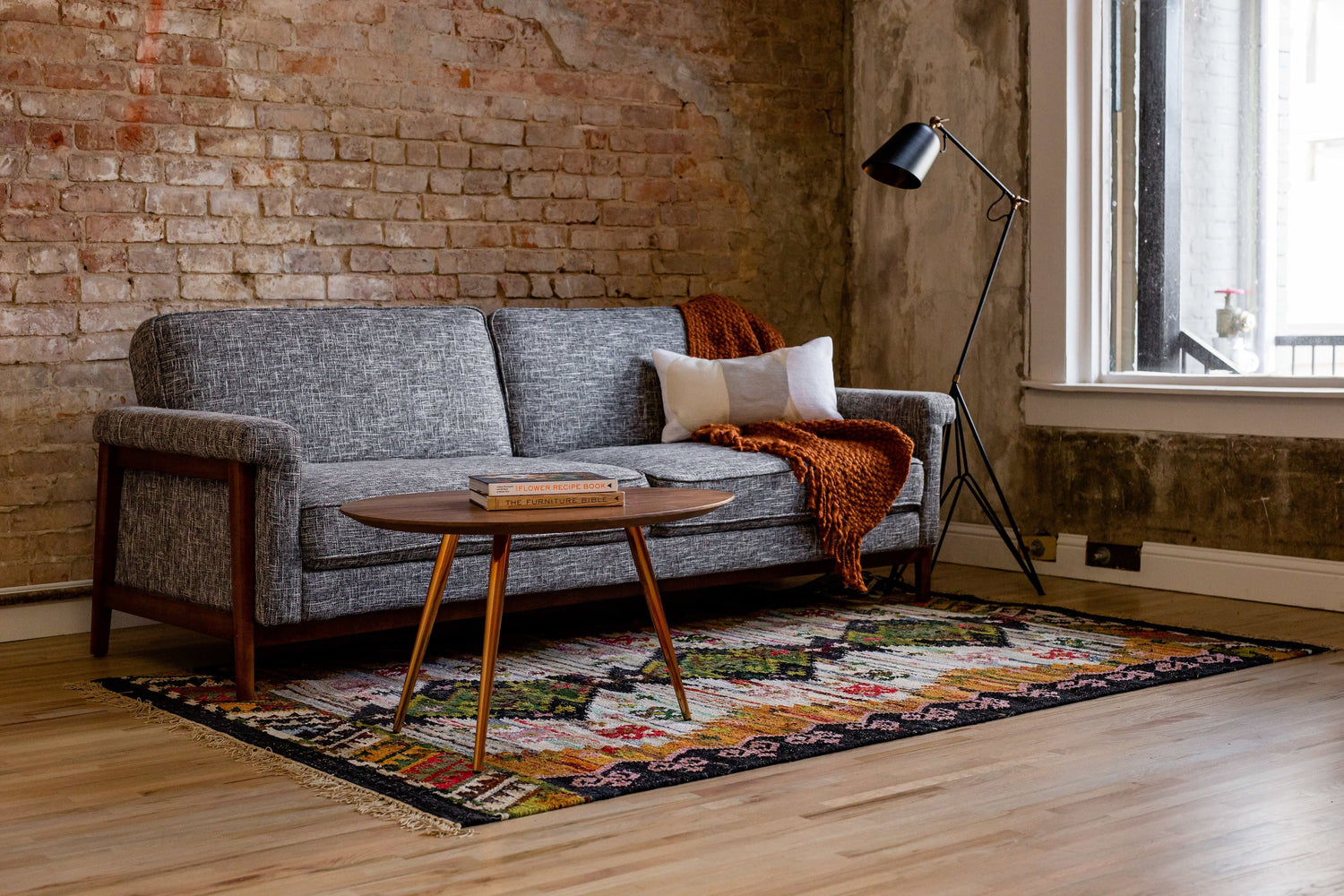 A cozy living room features a gray mid-century modern sofa with a white and orange throw blanket. In front is a wooden coffee table on a colorful patterned rug. The room has a rustic brick wall and large window, and is illuminated by a black floor lamp.