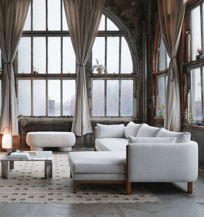 A stylish, industrial-style living room features large arched windows adorned with sheer curtains and an Emmy Sectional in light gray. A rectangular coffee table and ottoman rest on a patterned rug, while plants and a lamp bring warmth to the space.