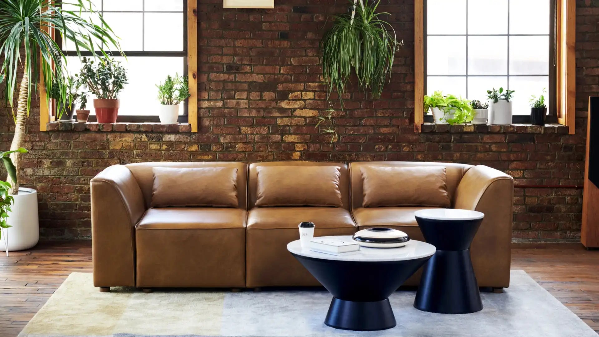 A modern living room showcases a Mambo Loveseat in brown leather positioned against a brick wall with windows. Potted plants decorate the windowsills. In front of the loveseat, there's a white round coffee table accompanied by two black side tables.