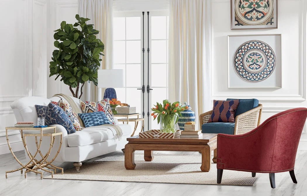 A bright living room with a white sofa adorned with colorful patterned pillows, a wooden coffee table, a red armchair, and a blue patterned chair. A large green plant stands beside the sofa. Decorative plates and wall art hang on white walls.