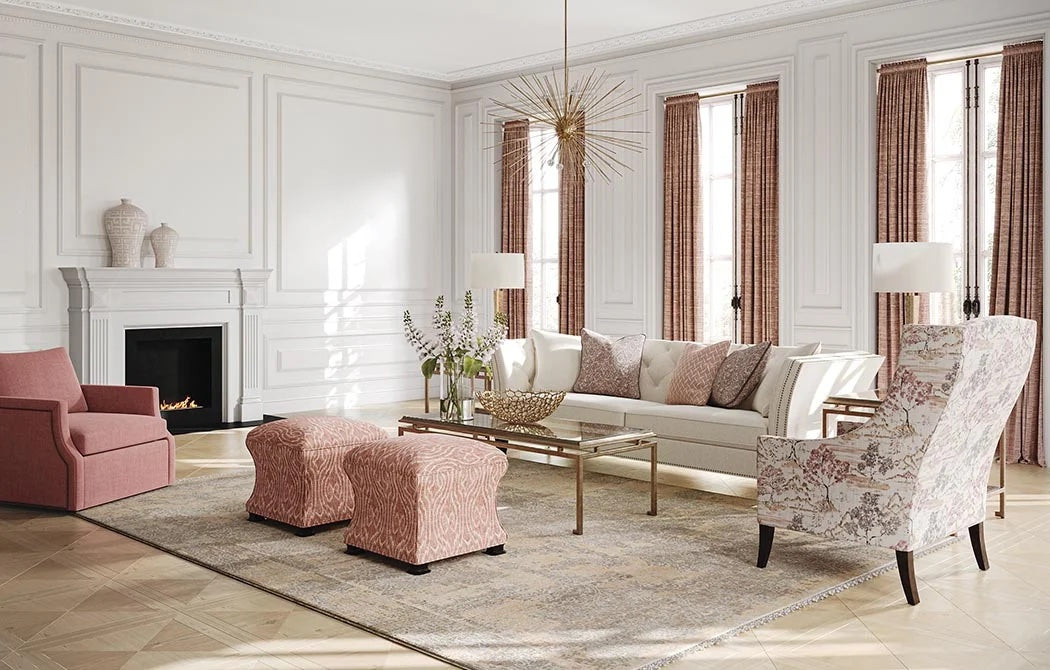 A sophisticated living room features a white sofa with patterned pillows, two pink ottomans, a patterned armchair, and a pink armchair. A gold coffee table and an elegant chandelier complement the white walls, tall windows with pink curtains, and a fireplace.