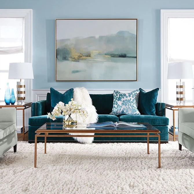 A modern living room with a teal velvet sofa adorned with blue and white pillows and a white throw blanket. A glass coffee table with a book and decorative items sits on a plush white rug. The walls are light blue, featuring a large abstract painting.