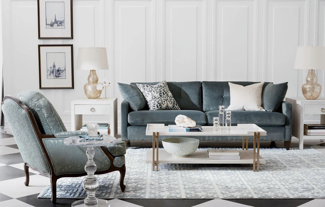 A stylish, modern living room featuring a plush teal sofa with decorative pillows, a light blue armchair with a clear side table, two white side tables with lamps, and a central white coffee table with decor items. The room has a checkered floor, white walls, and framed art.