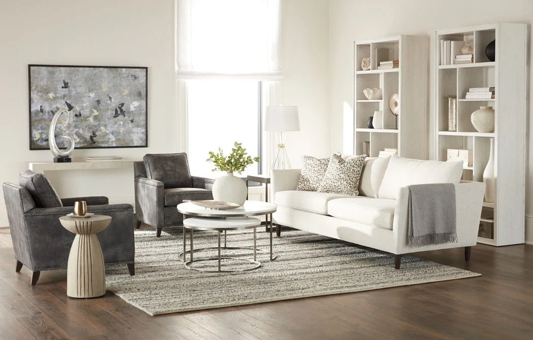 A modern living room with a white sofa, grey armchairs, and a round coffee table on a striped rug. The room features a large abstract painting, two tall bookshelves with decor items, and a floor lamp. A small side table with a vase and greenery adds a touch of nature.