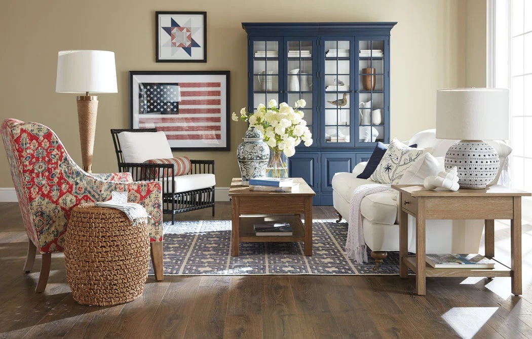 A cozy living room with a white sofa, a patterned armchair, a wicker basket, and a wooden side table. A blue china cabinet and a framed American flag wall art add character. The room is brightly lit by a large window on the right. There's a modern rug on the wooden floor.