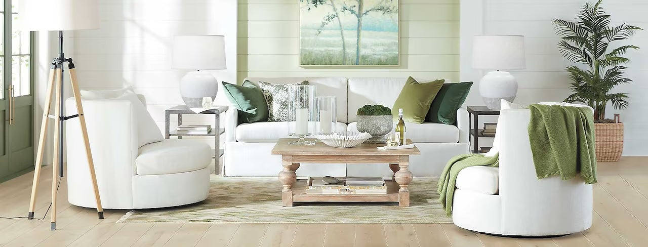 A bright living room with white walls and hardwood flooring features a white sofa adorned with green and white pillows, flanked by two white armchairs. A wooden coffee table is in the center, and a large painting of trees hangs above the sofa. A plant is in the right corner.