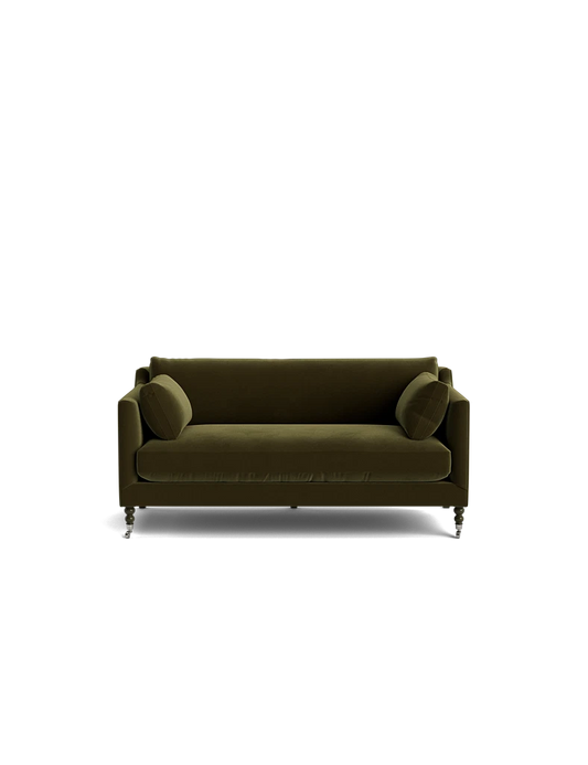 A Fabienne Sofa in green velvet, complete with two cylindrical cushions, set against a plain black background. The sofa boasts a modern design with clean lines and is supported by small casters.