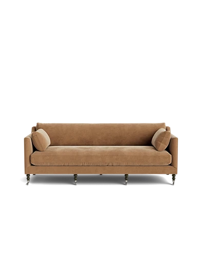 The Fabienne Sofa, upholstered in beige velvet and accompanied by two matching cylindrical pillows, rests on six wooden legs set against a black background, showcasing its simple, modern design with clean lines.