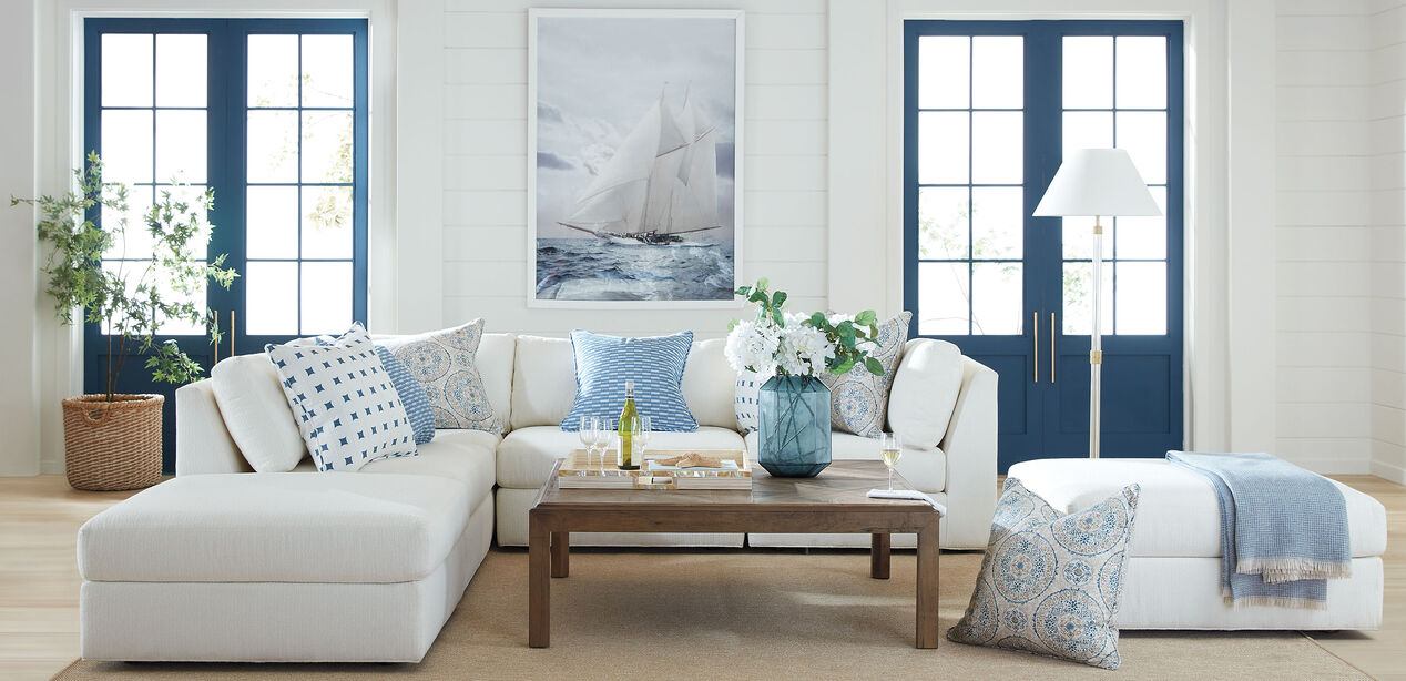 A bright living room featuring the Daydreamer Modular Six-Piece Sectional adorned with patterned blue and white pillows, a wooden coffee table with decor, and a nautical painting above. The space also includes large windows with blue frames, a potted plant, and a floor lamp.