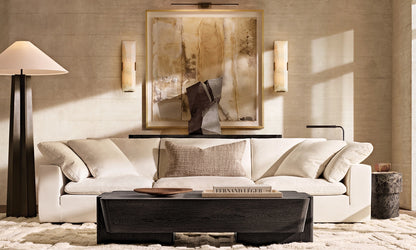 A contemporary living room showcases the Cloud Modular 3-Piece Sofa adorned with cushions, accompanied by a dark wooden coffee table and an oversized abstract painting on the wall. Flanking the artwork are two wall sconces, with a floor lamp positioned to the left, establishing a cozy and stylish atmosphere.