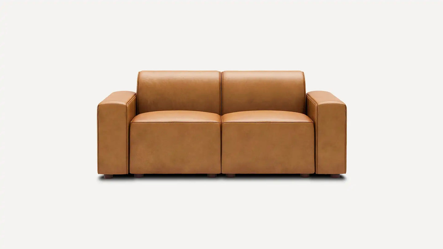 The Mambo Loveseat, featuring a modern design with tan leather upholstery and spacious, blocky armrests, is centered against a plain white background.