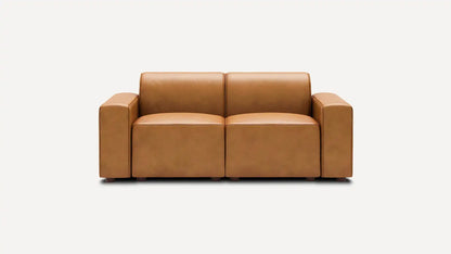 The Mambo Loveseat, featuring a modern design with tan leather upholstery and spacious, blocky armrests, is centered against a plain white background.