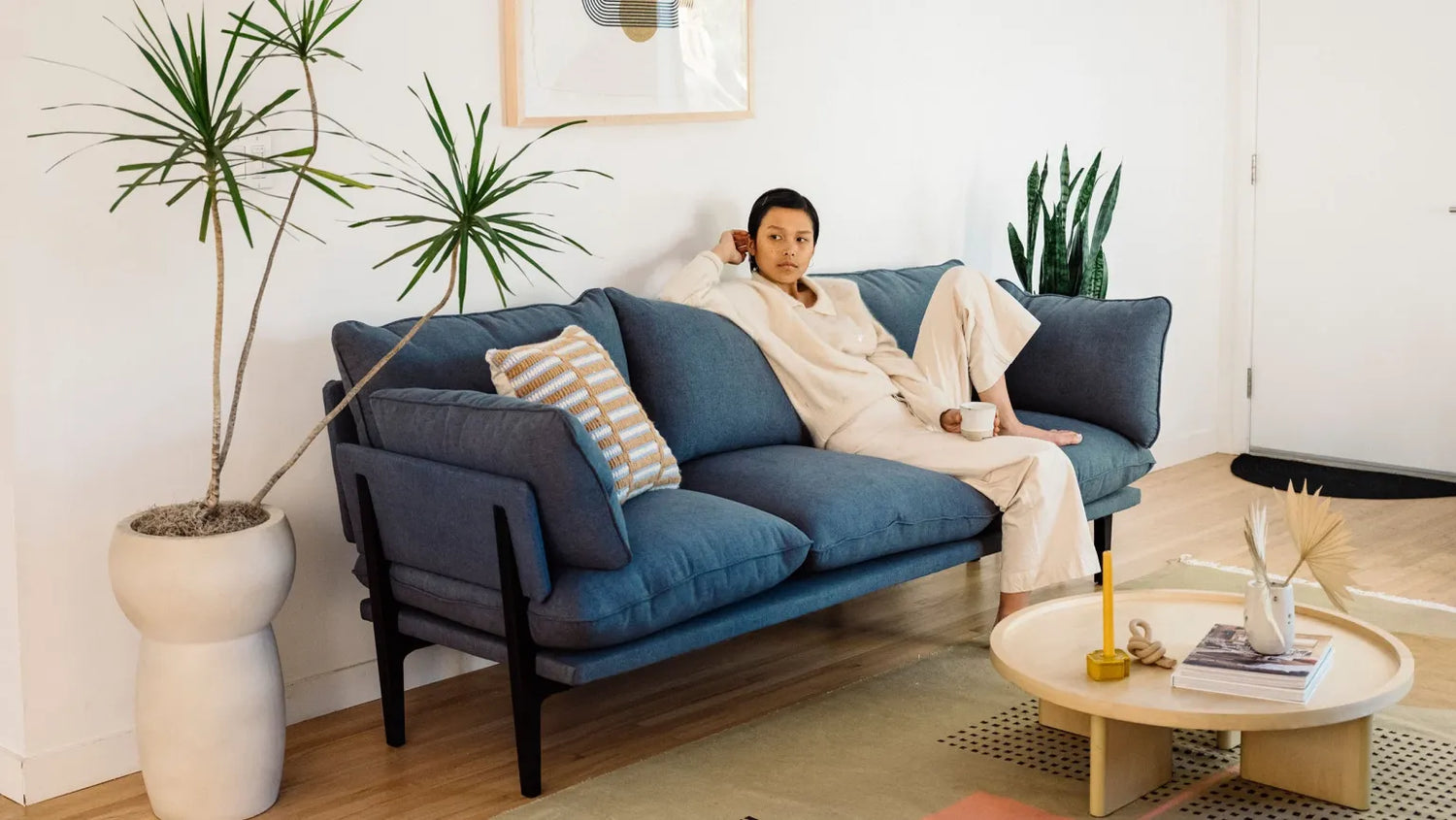 A person lounges on a blue sofa in a modern living room. They are holding a mug and are dressed in light-colored clothes. The room features minimalistic decor with potted plants, a round coffee table with books and candles, and a framed picture on the wall.