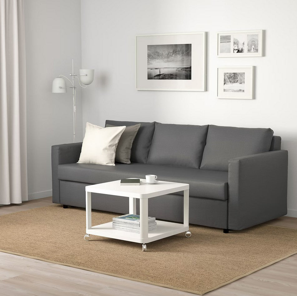 A modern living room with a FRIHETEN Sleeper Sofa, paired with a white coffee table on a beige rug, and framed black-and-white art adorning the walls. A floor lamp is strategically placed next to the sofa, complemented by a white curtain hanging beside it.