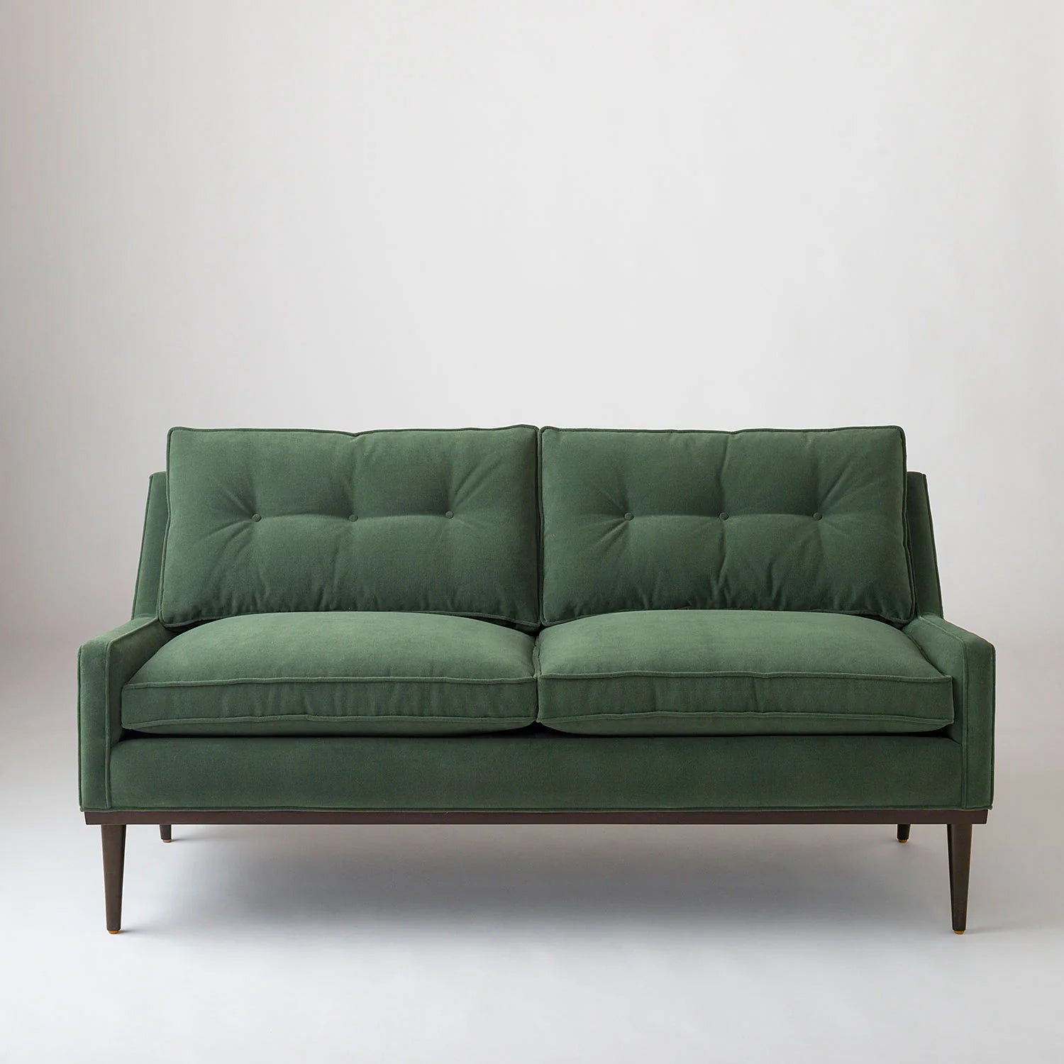 The Jack Sofa, featuring a sleek, minimalist design with tufted cushions and wooden legs, is positioned against a plain white background. This mid-century modern piece comes in a green color.