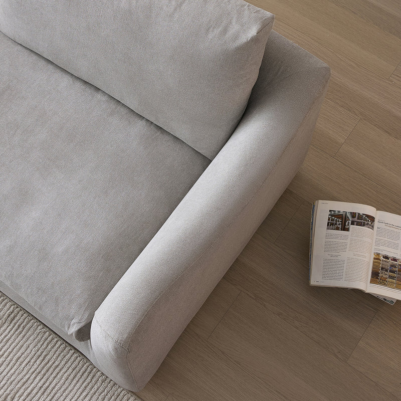 A top view of the Feathers 88" Left Facing Sectional in gray, featuring a cushioned back and armrest, is positioned on a wooden floor next to an open magazine. A beige rug is partially visible in the lower left corner.