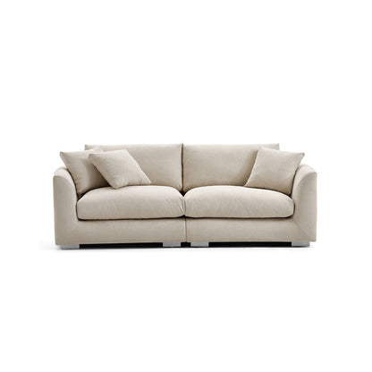 The Feathers 3-Seater Sofa, in a cream color, boasts a contemporary design with wide cushions and four matching throw pillows. It is displayed against a white background, highlighting its simple yet elegant style.