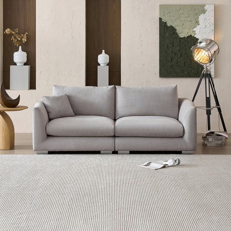 A modern living room with the Feathers 3-Seater Sofa in gray, accompanied by a small pillow and a magazine on a textured rug. The wall features two vases on shelves and an abstract art piece. A large, vintage-style floor lamp stands to the right.