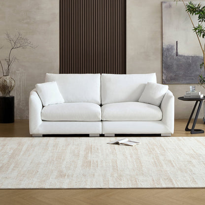 A modern living room featuring the Feathers 3-Seater Sofa in white, adorned with two cushions, is set against a neutral wall with minimalistic decor. A small side table holds a plant and book, while a large rug covers the wooden floor. Abstract art and potted plants add elegance to the space.