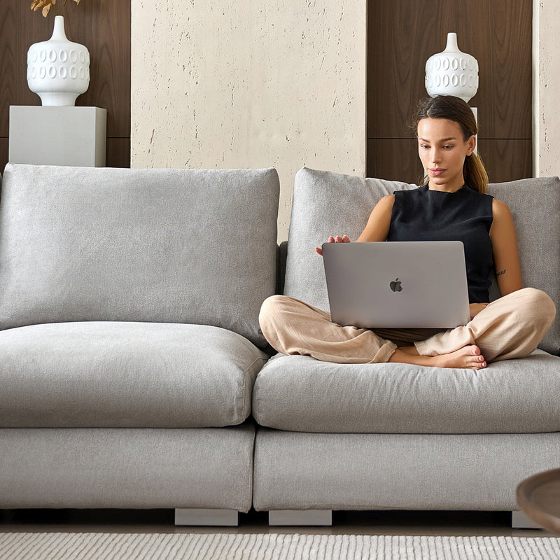 A woman sits cross-legged on a Feathers 3-Seater Sofa, working on a laptop. She is wearing a black sleeveless top and beige pants. The background features a textured wall with two decorative vases, creating the ambiance of a modern, cozy living room.