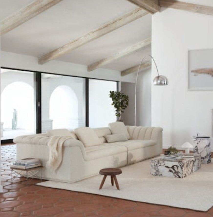 In a contemporary living room, a beige Float Sofa is accompanied by a marble coffee table on a terracotta tile floor. A tall arched lamp stands next to a potted plant in the corner, with large windows allowing natural light to stream in. The ceiling features wooden beams.