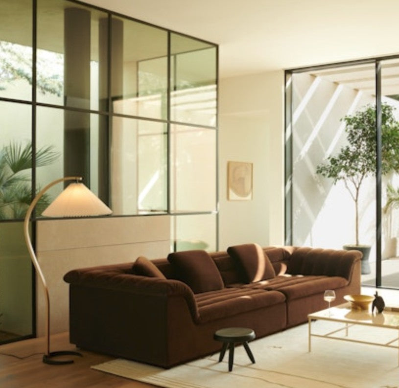 A modern living room featuring the Float Sofa upholstered in brown velvet, complemented by a curved floor lamp, a small round stool, and a glass coffee table. Large windows showcase leafy plants outside, while natural light illuminates the space.