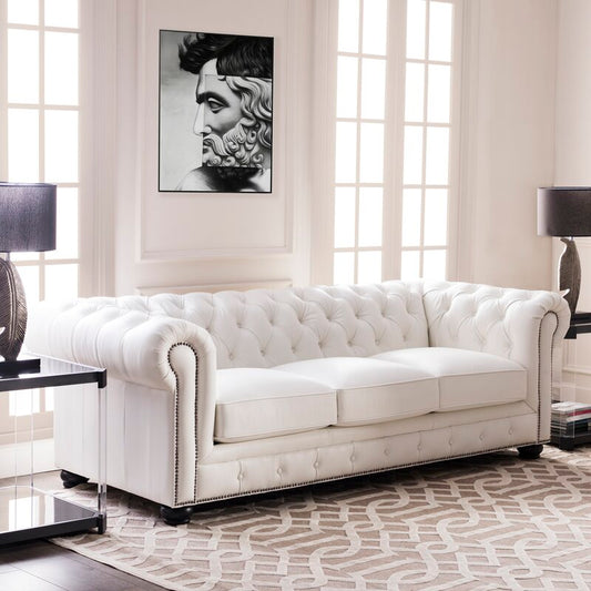 A Birch Lane Freddie 95'' Leather Sofa sits in a bright living room with large windows. A black and white artwork of a face hangs above, while two black lamps flank the sofa, and a patterned rug adds contrast to the floor.