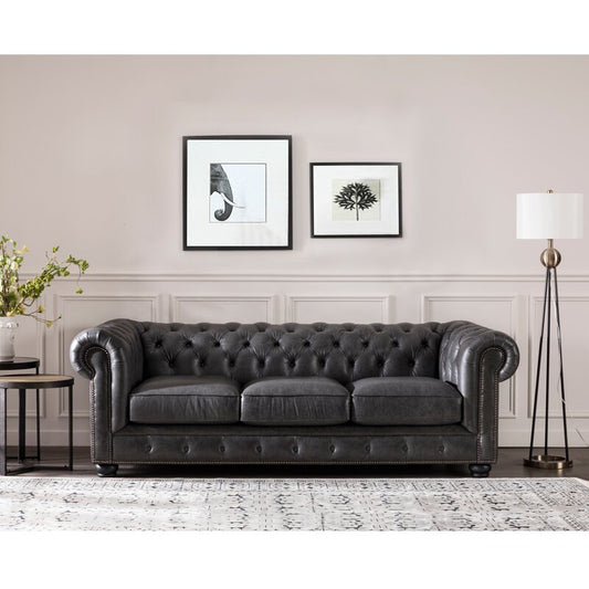 A luxurious Birch Lane Freddie 95'' black leather sofa in a modern living room, flanked by a side table with a vase of flowers and a tall floor lamp. Two framed artworks hang on the wall above, while a patterned rug covers the floor.