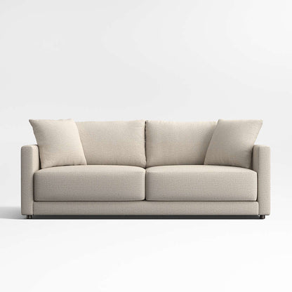 The Gather Sofa 89" is a beige, modern piece featuring straight arms and two large seat cushions. Each end of the sofa is accentuated with matching throw pillows. Set against a plain white wall and floor, the simplistic, contemporary design of the sofa stands out.