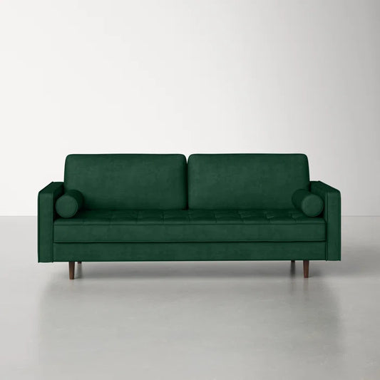 The Geo 84'' Upholstered Sofa, featuring a modern design with green velvet upholstery, includes two seat cushions and two cylindrical bolster pillows. It stands in an empty room with a light-colored floor and wall, supported by four wooden legs that enhance its minimalist aesthetic.