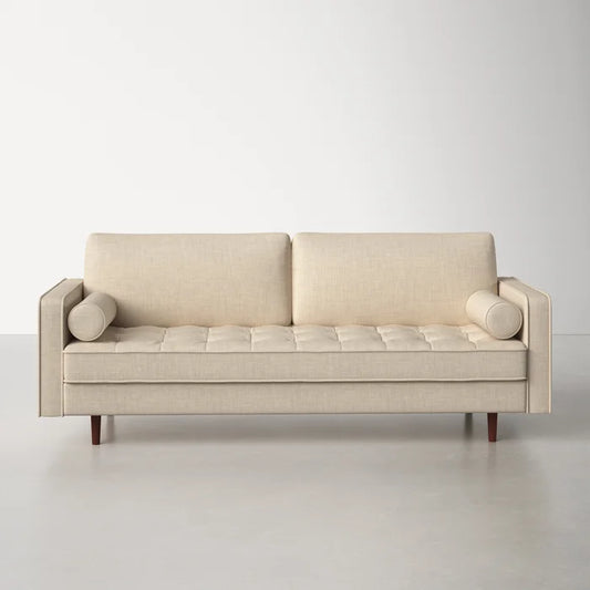 The Geo 84'' Upholstered Sofa, featuring a beige modern design with clean lines and tufted seat cushions, includes two back cushions and two cylindrical side pillows. It rests on four wooden legs and is placed against a plain white wall on a smooth, light gray floor.