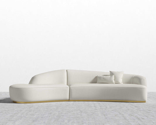 The Reya Curved Sectional is a sleek, modern sofa with soft lines and a minimalist design, featuring three matching cushions. It is displayed against a plain white wall on a light gray textured floor.