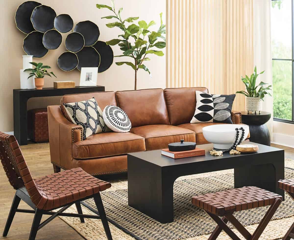 A modern living room features a brown leather sofa adorned with patterned pillows, a black coffee table with decorative items, woven chairs, and lush green plants. A textured rug anchors the space, and abstract black wall art adds a stylish touch.