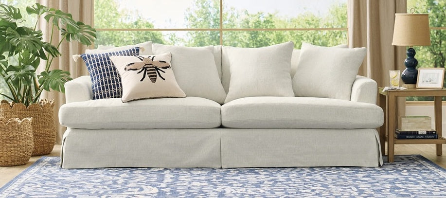 A cozy living room featuring a light-colored sofa with multiple white cushions and a decorative pillow with a bee design. The sofa sits on a blue patterned rug. A wooden side table with a lamp and books is next to the sofa, and large windows reveal greenery outside.