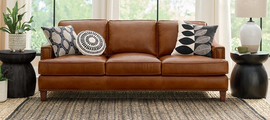 A brown leather sofa with nailhead trim sits in a well-lit living room with large windows. The sofa has four decorative pillows with black and white geometric patterns. To the left is a potted plant on a small black side table; to the right, a lamp on another similar table. The floor features a woven rug.