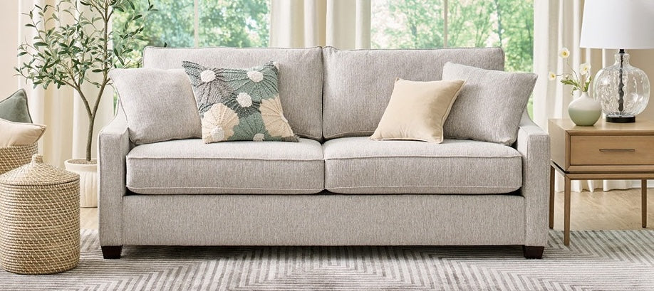 A modern living room features a light gray sofa adorned with patterned and plain cushioned pillows. A wooden side table with a decorative lamp and vase sits nearby. A woven pouf is on one side, and large windows with curtains reveal greenery outside.